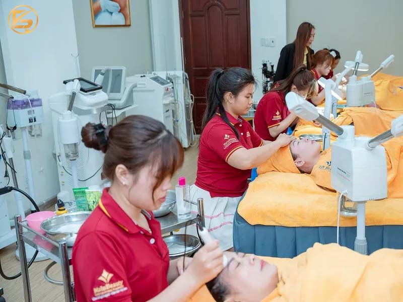 Học viên MainBeauty mặc đồng phục đỏ đô đang thực hành chăm sóc da và đi máy thẩm mỹ trên mẫu thật.